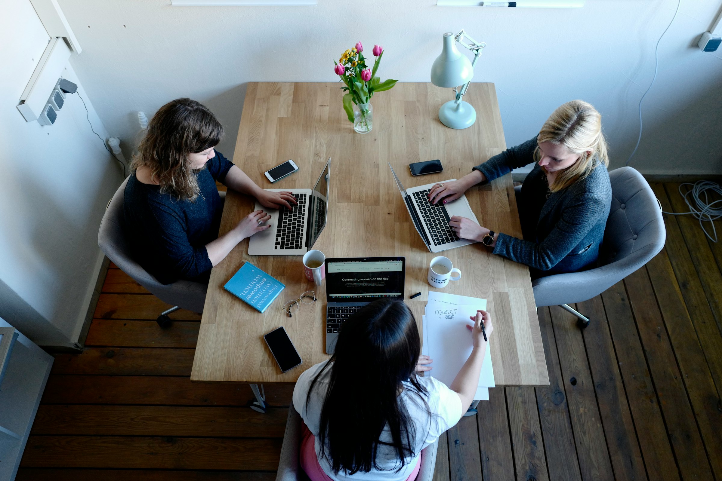 Tres personas trabajando con portátiles alrededor de una mesa de madera, vistas desde arriba
